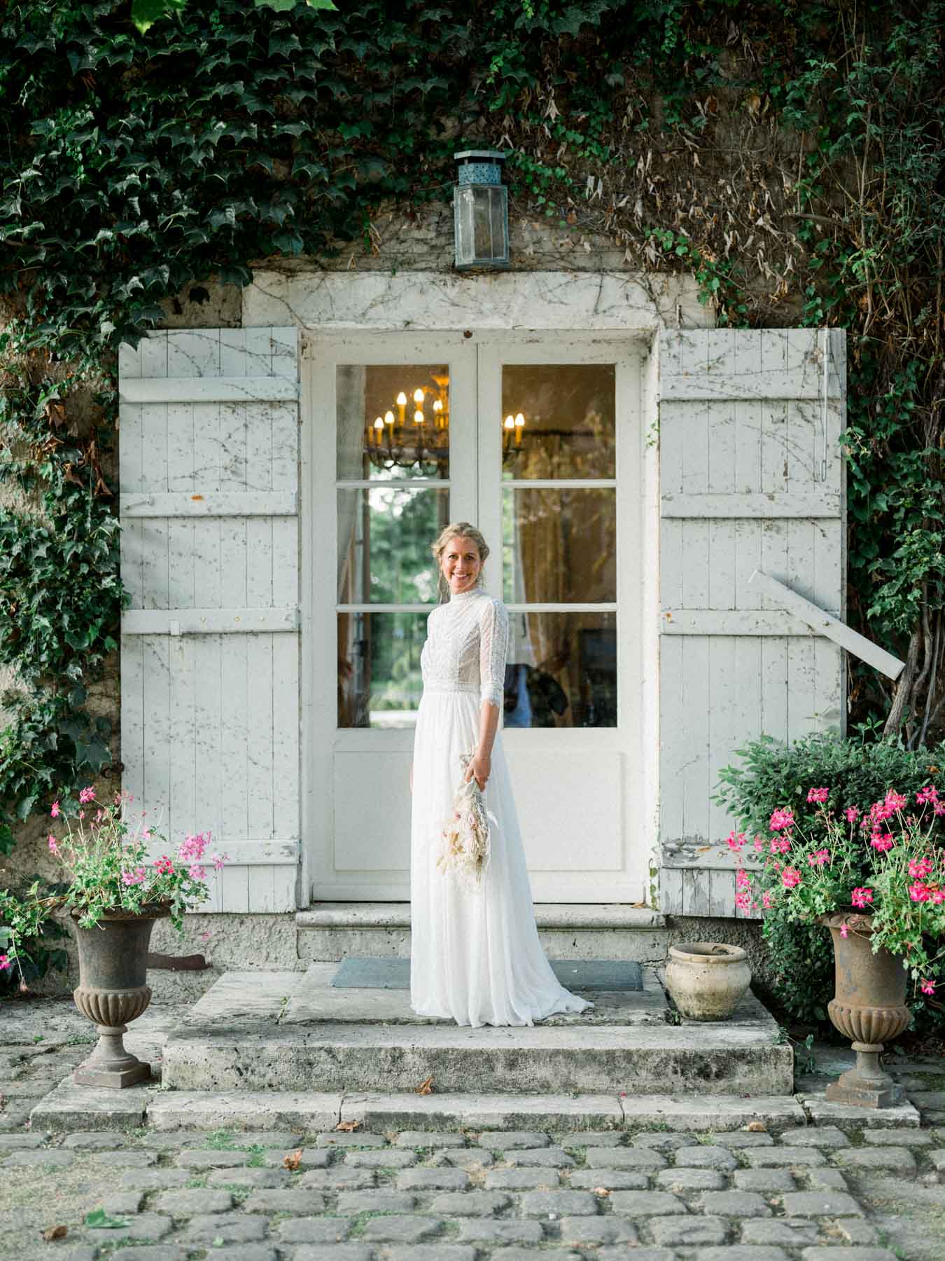 The bride in a lace gown on the steps of a door with white shutters