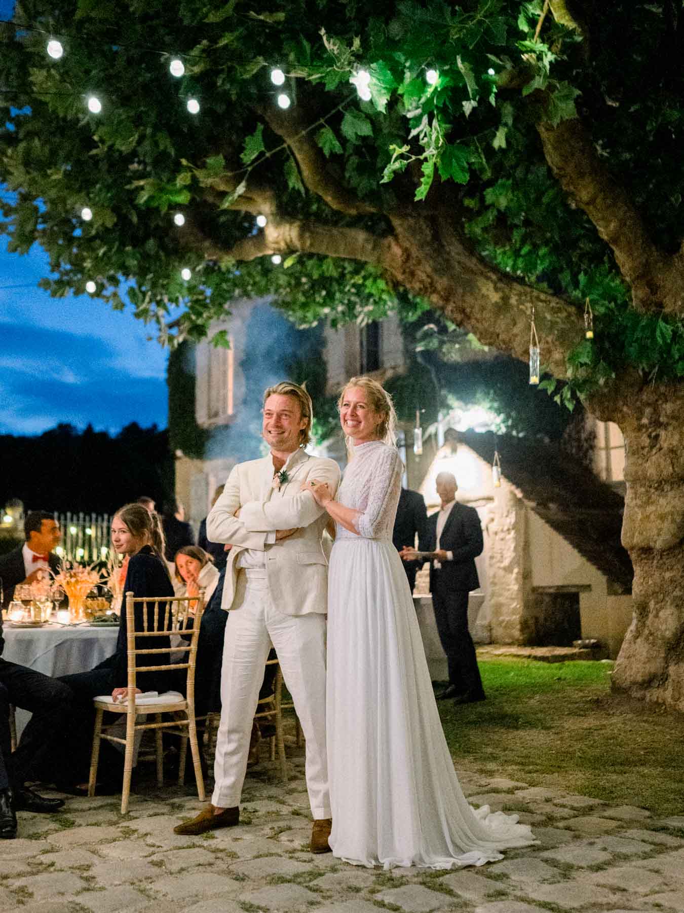 The couple standing during dinner beneath trees strung with lights