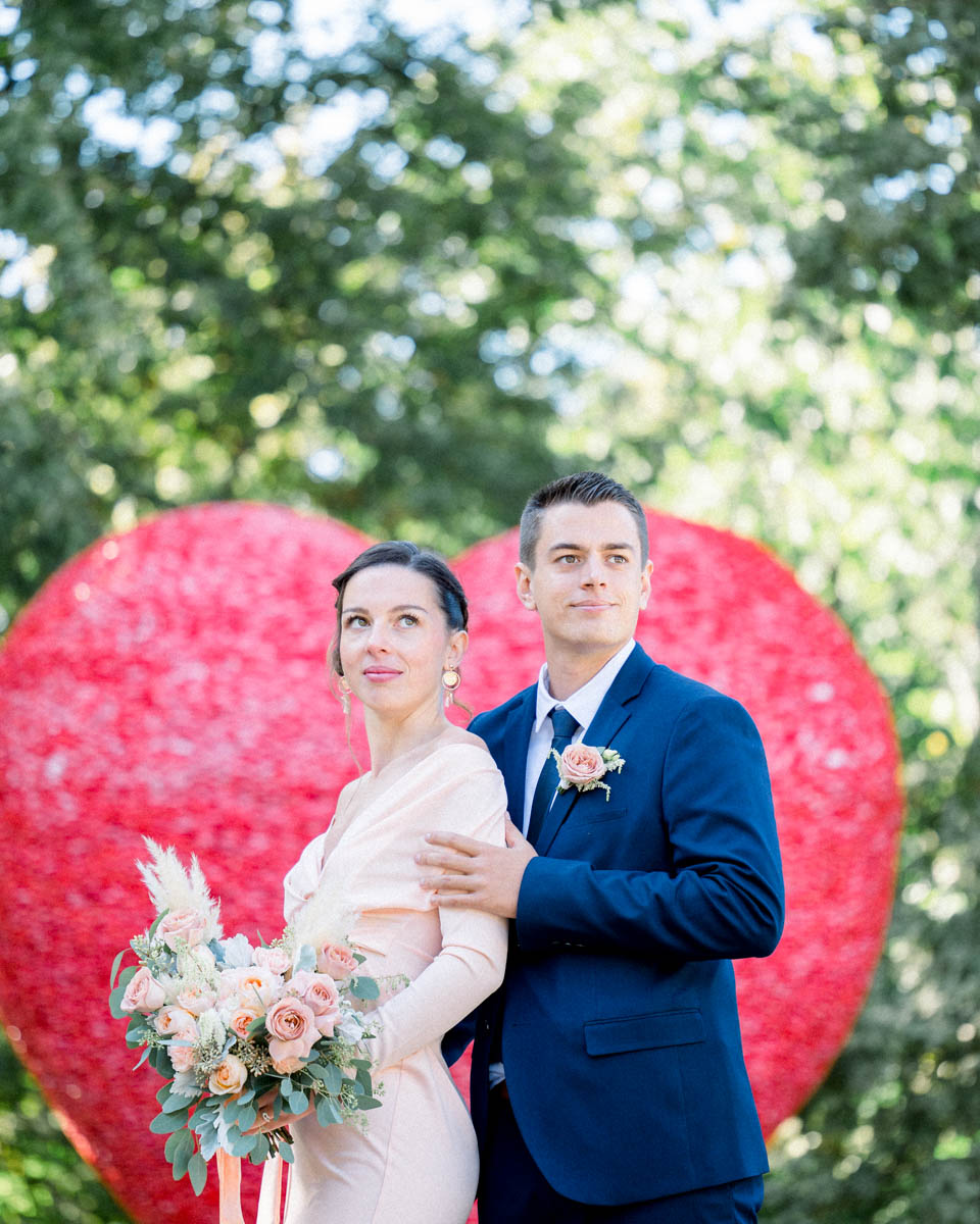 The couple posing together in front of the giant red heart, bouquet in hand