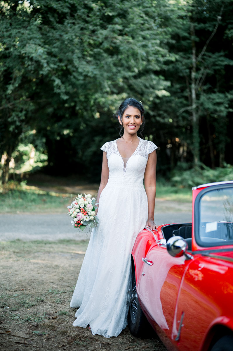 The bride with her bouquet leaning on the red convertible in the forest