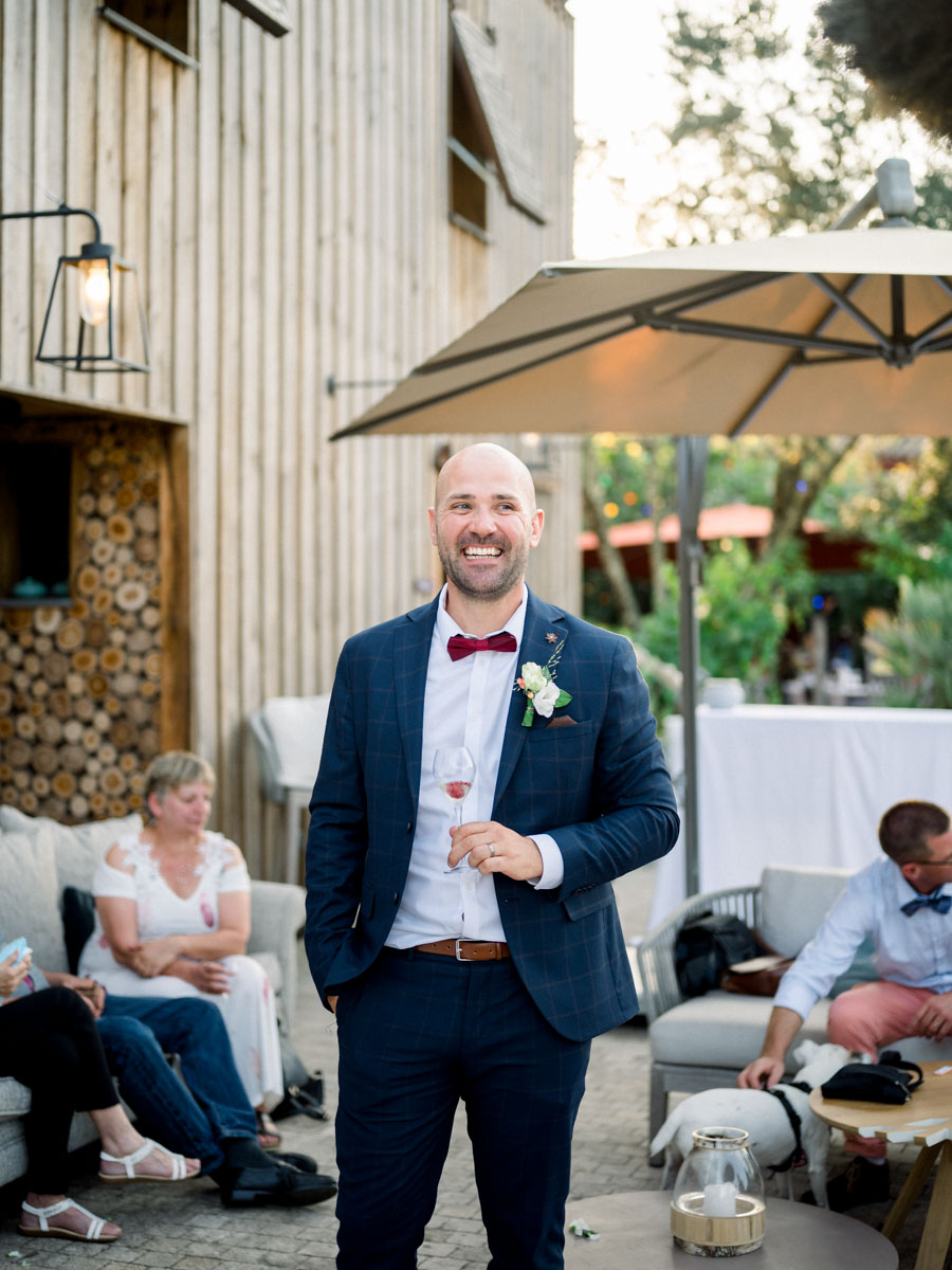 The groom in a blue suit and red bow tie, glass in hand, at the drinks