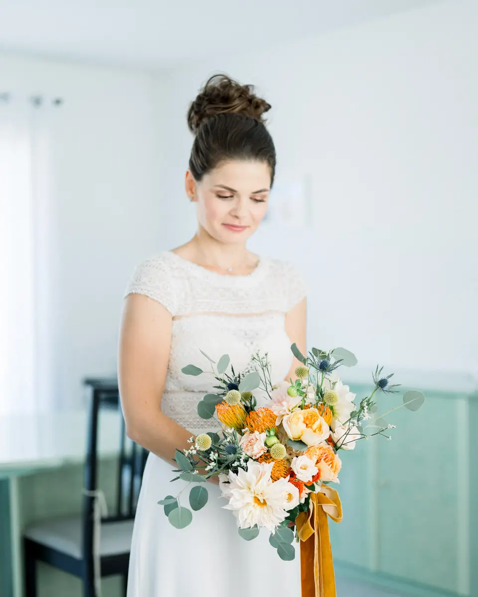 The bride looking down at her orange and white bouquet with ochre ribbons