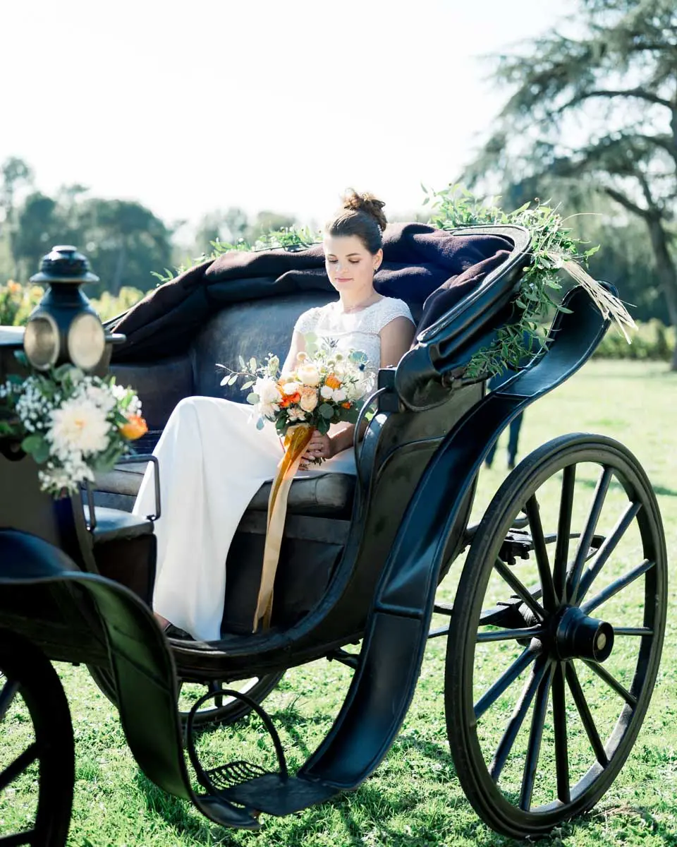 The bride seated in a black horse-drawn carriage with an orange bouquet