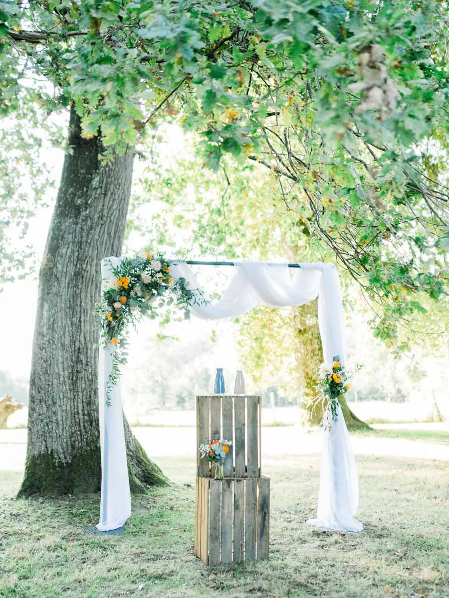 White draped arch with orange flowers at the foot of a large tree