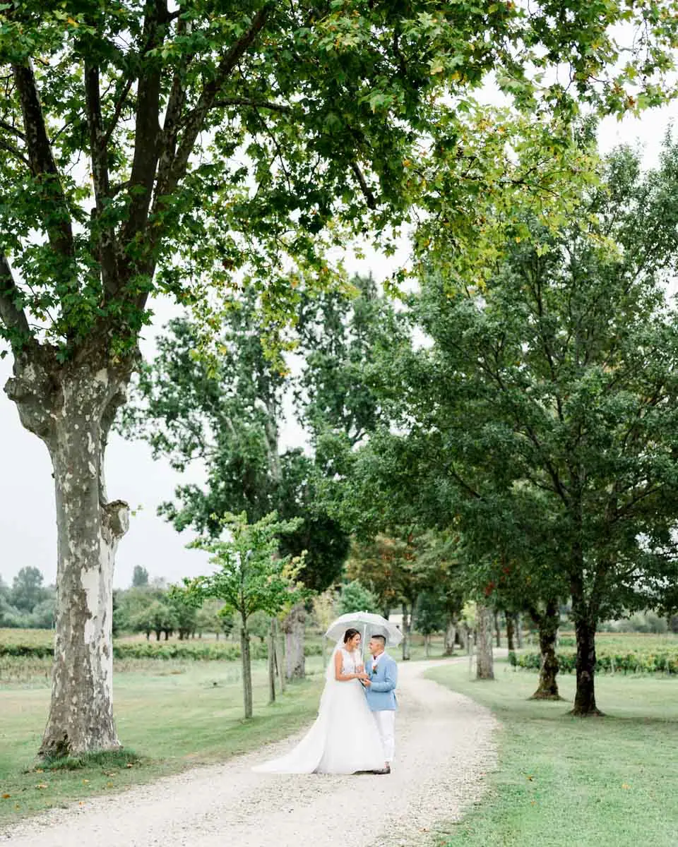Bride and groom under an umbrella on an avenue of tall trees on the estate
