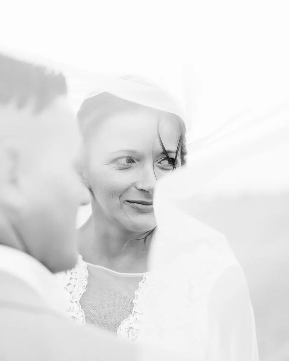 Bride smiling beneath her veil beside her partner, in black and white