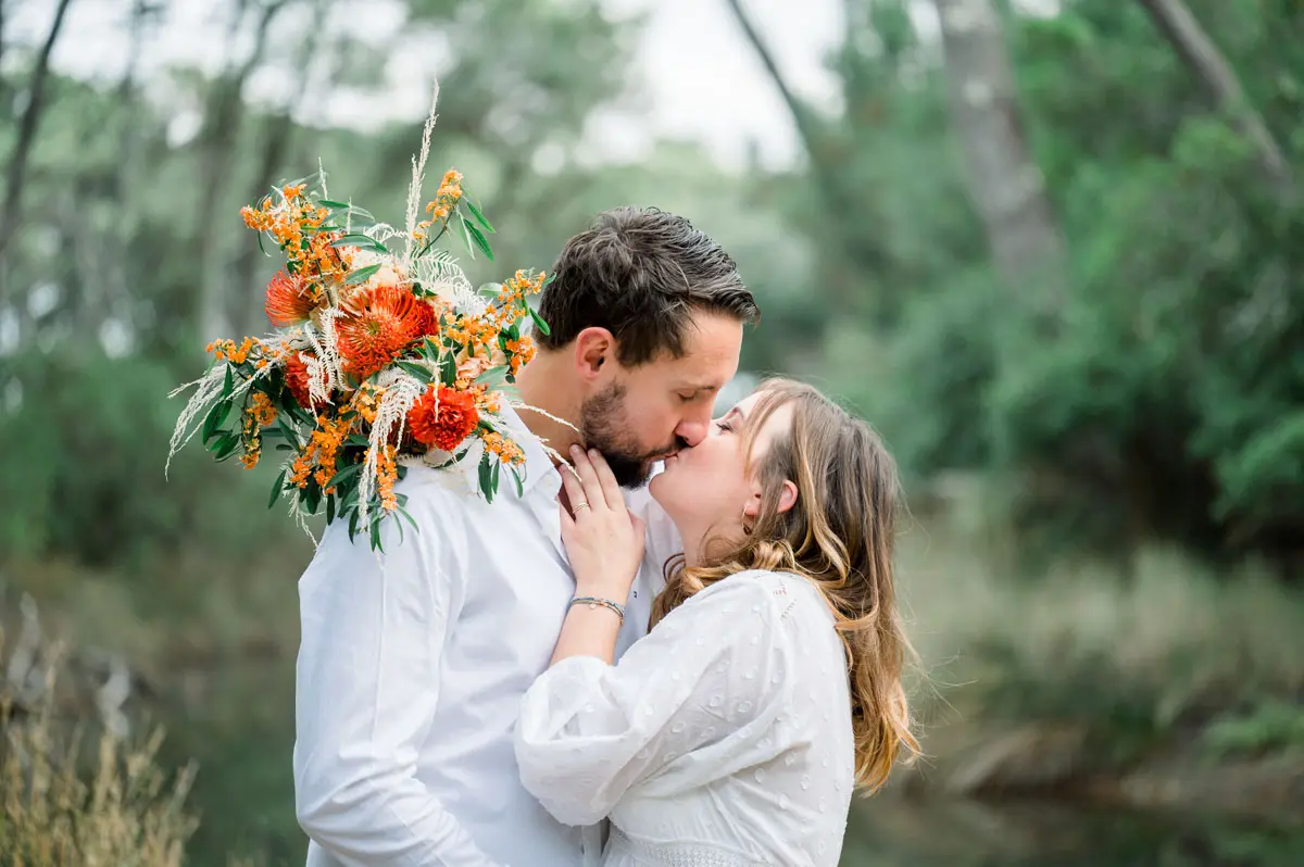 The dad-to-be kisses his pregnant partner, orange bouquet raised, in the forest