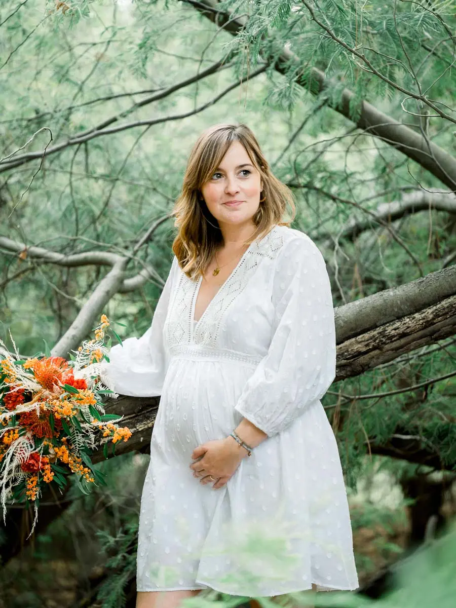 Mum-to-be in a white dress perched on a low branch, bouquet in hand