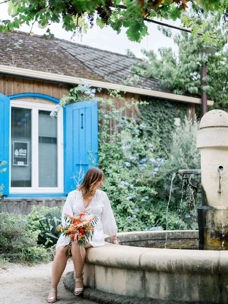 Mum-to-be sitting on a stone fountain by a cabin with blue shutters