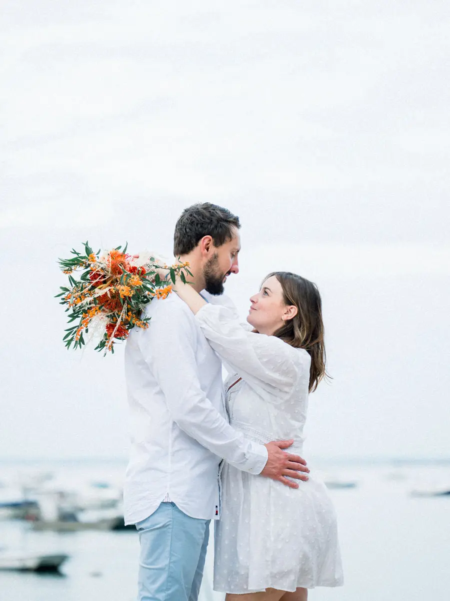 The parents-to-be embracing at the water's edge, orange bouquet in hand