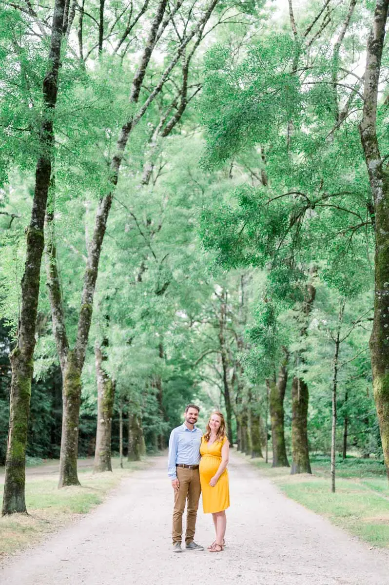 Expectant couple walking hand in hand down a tree-lined avenue in Parc Bordelais
