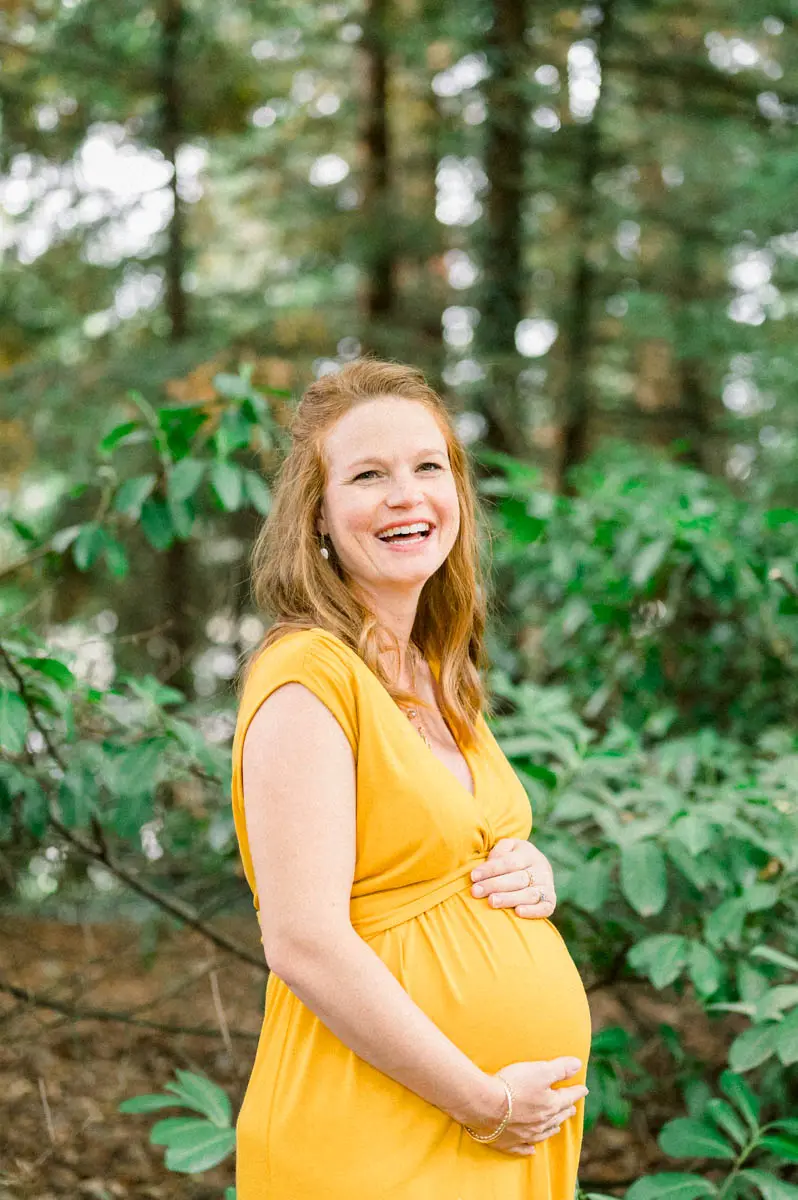 Mum-to-be in a yellow dress laughing with her hands on her bump among the trees