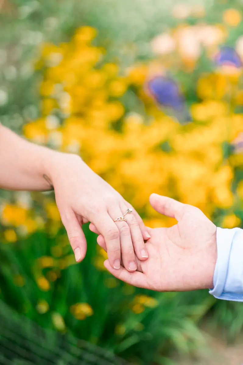 The couple's intertwined hands in front of a bed of yellow flowers