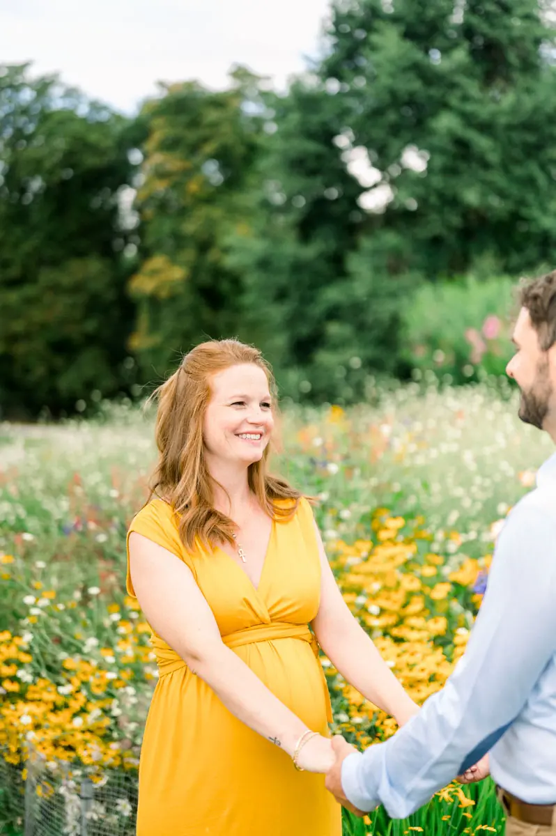 Mum-to-be in a yellow dress holding her partner's hand by a flower bed