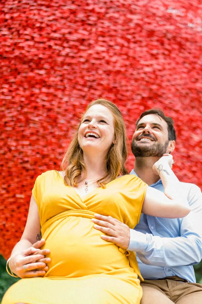 Expectant couple laughing against a red wall, his hand on her bump