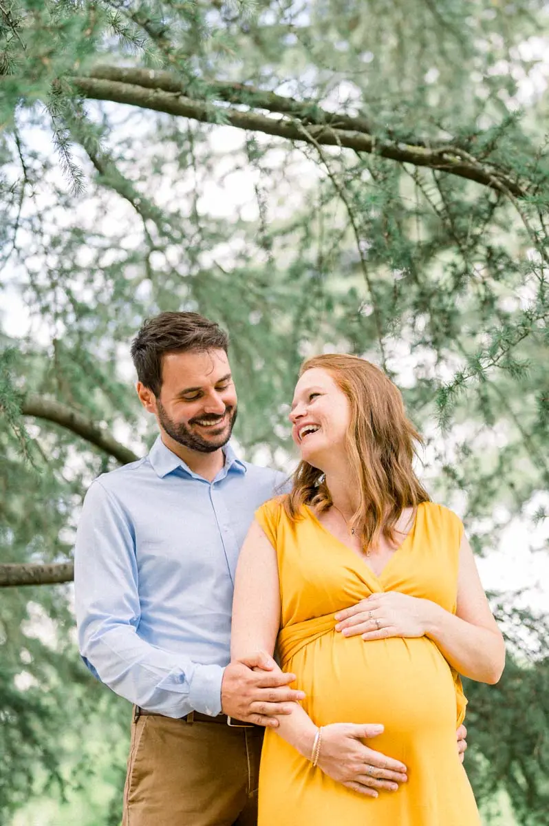 The dad-to-be holds his pregnant partner in a yellow dress beneath a cedar