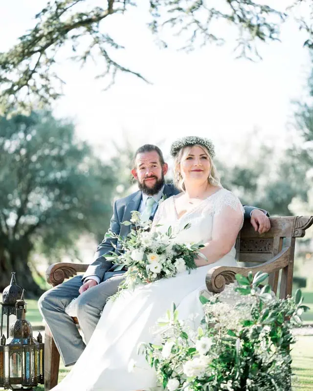 The couple seated on a bench during the outdoor ceremony, surrounded by flowers