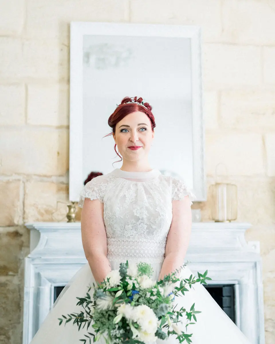 The bride in a lace gown holding her white bouquet by a fireplace