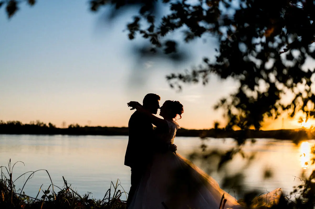 The couple silhouetted by the water, framed by branches at dusk