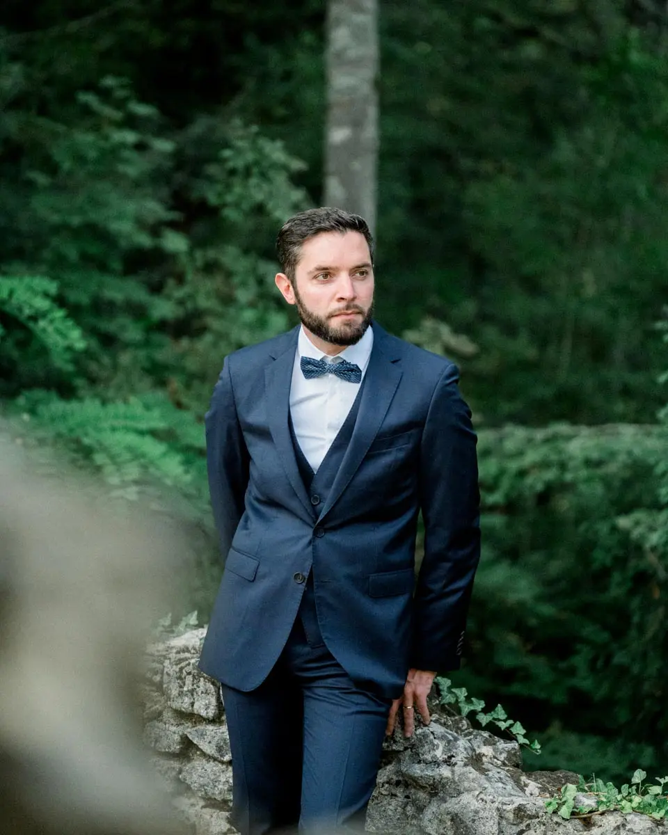 Groom in a navy suit leaning against a rock in Parc Majolan