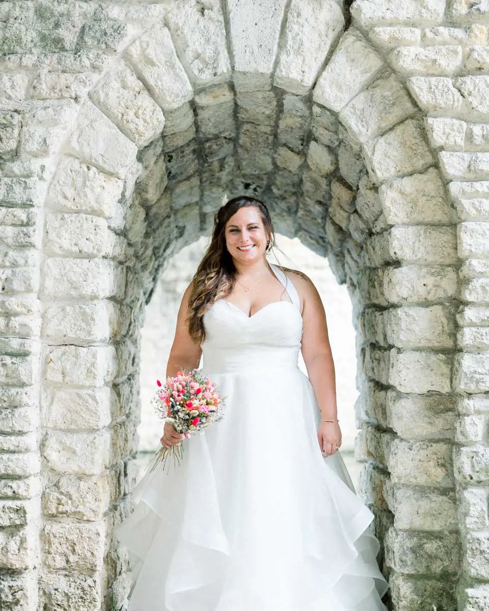 Bride standing beneath a stone arch in Parc Majolan, holding her bouquet