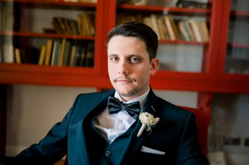 Groom in a midnight blue suit and bow tie in front of a red bookcase