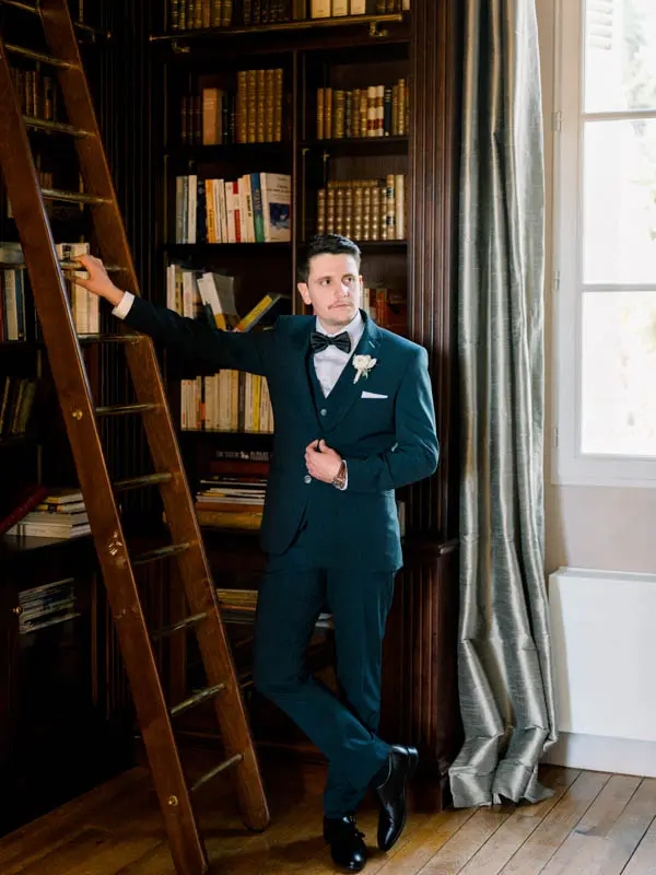 Groom leaning against a wooden library bookcase with a ladder at Villa La Tosca