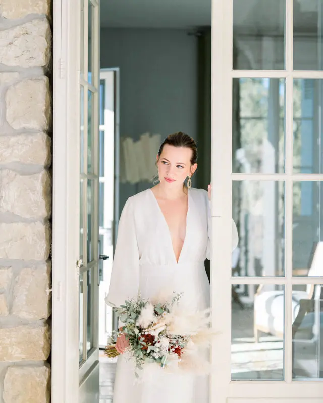 Bride in a white dress stepping between the French windows with her bouquet