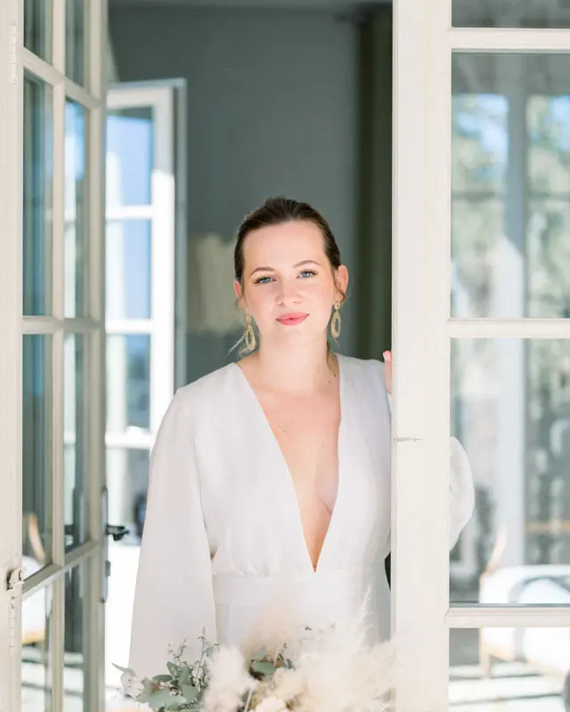 Smiling bride leaning on a French window, pampas bouquet in hand