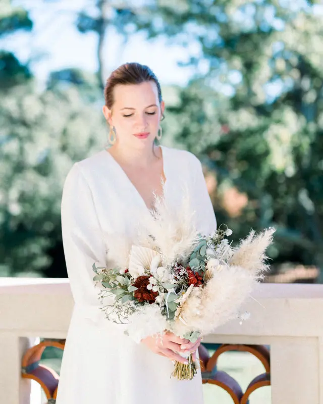 Bride gazing at her dried flower bouquet on the balcony, pines behind her
