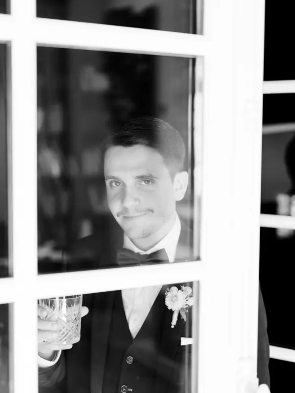 Groom waiting behind a window, glass in hand, in black and white
