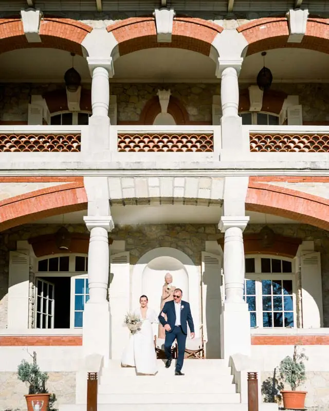 Romane and Julien walking out of the villa beneath its arches after the ceremony