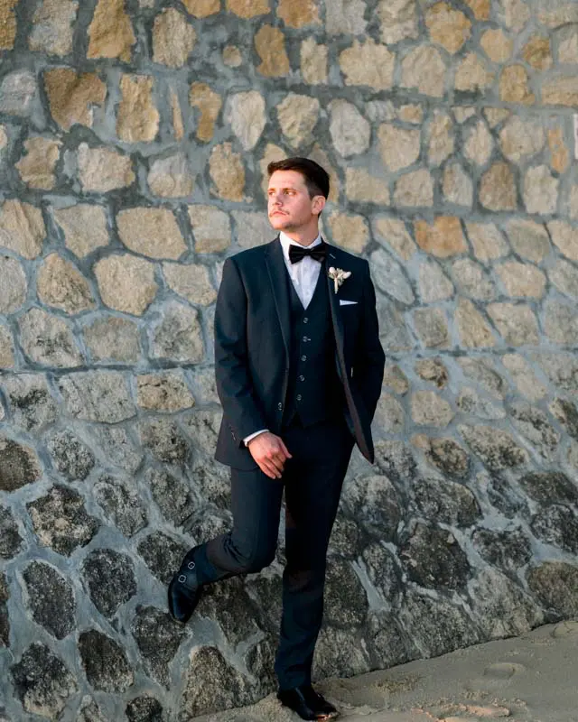 Groom in a three-piece suit leaning against a stone wall
