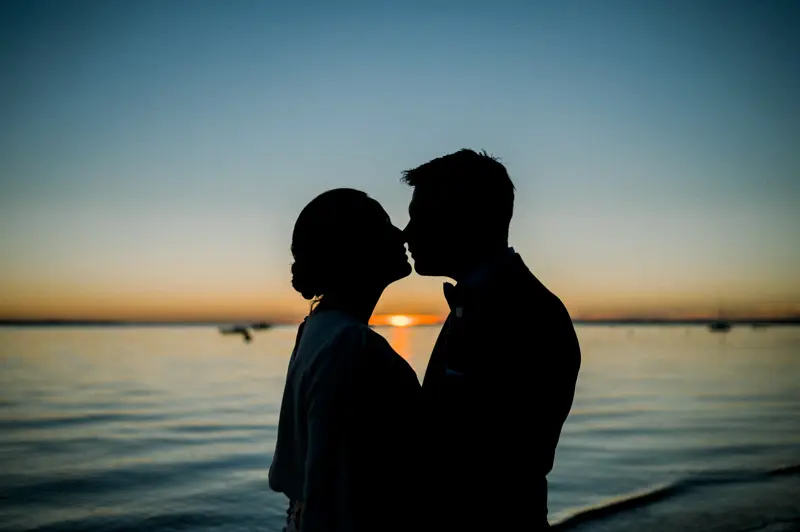 Silhouettes of the bride and groom kissing against the sunset over the water