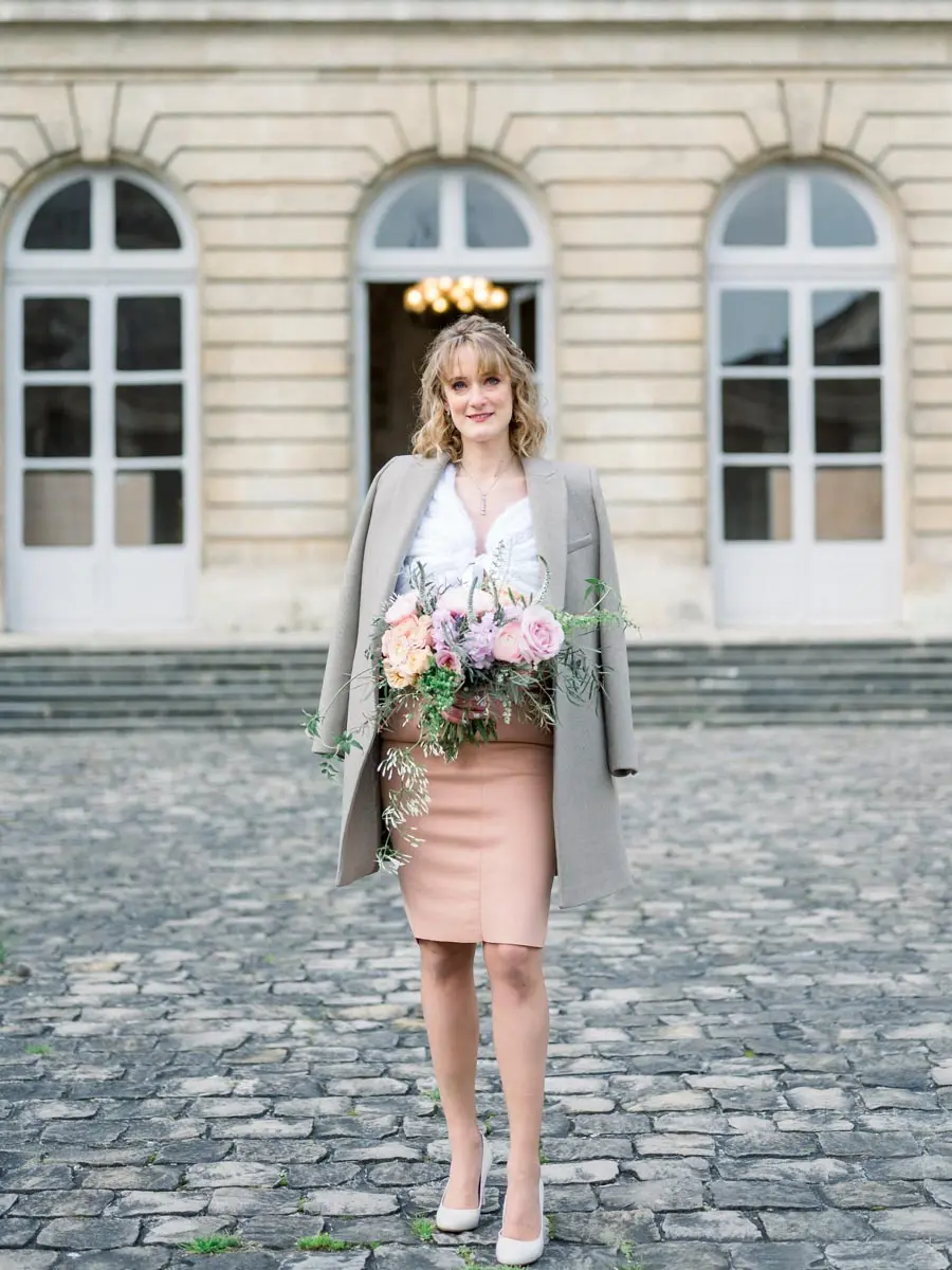 Marjorie in a grey coat and pink skirt holding her bouquet in the town hall courtyard