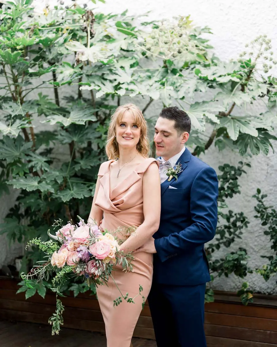 Bride in a dusty pink dress and groom in a blue suit before a wall of greenery