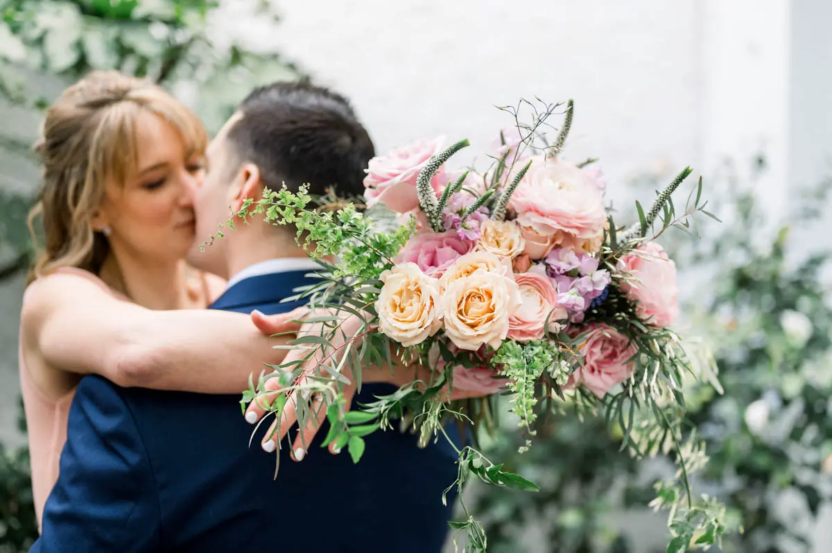 The couple kissing behind a bouquet of pink and peach flowers