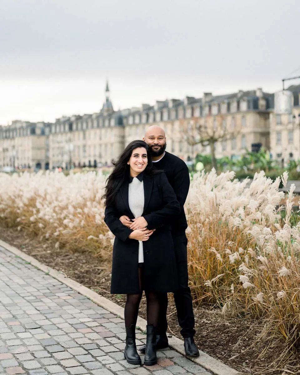Engaged couple embracing among ornamental grasses before Bordeaux's riverside facades