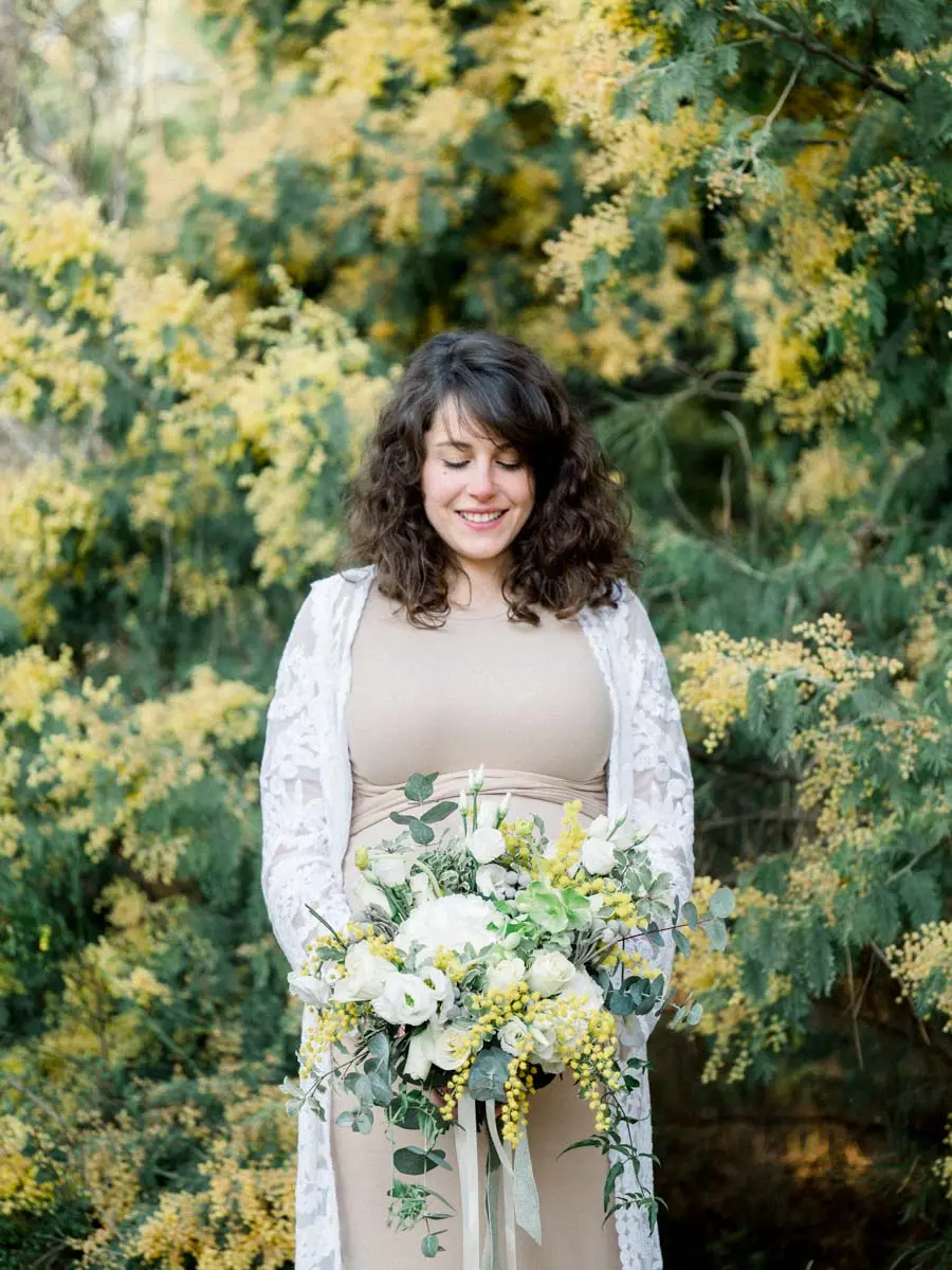 Mother-to-be in a beige dress and lace cardigan before a mimosa tree