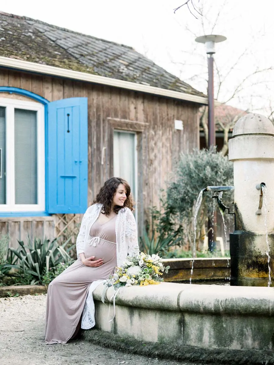 Mother-to-be sitting on a fountain by a wooden cabin in the village of L'Herbe