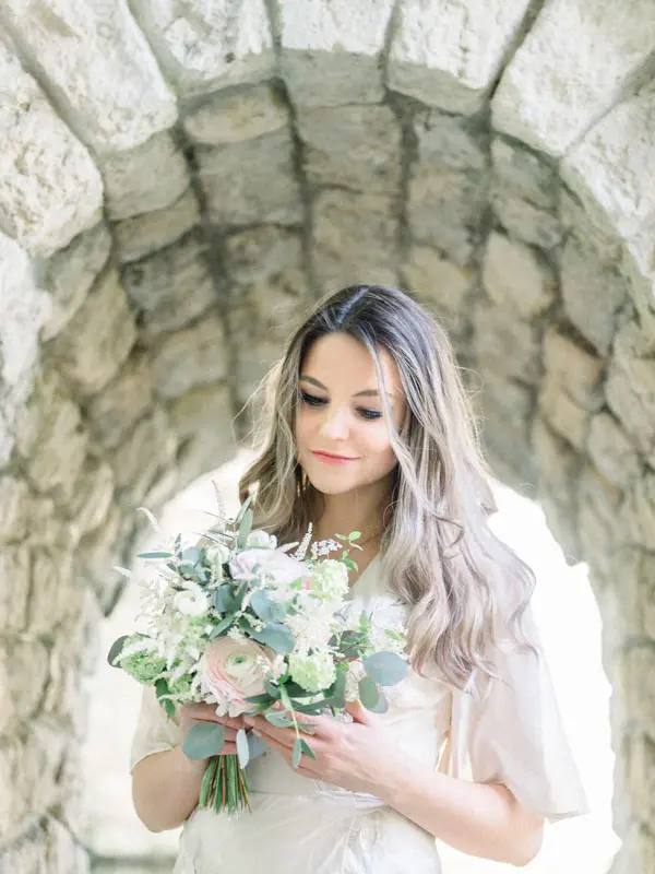 Bride-to-be beneath a stone arch, looking down at her bouquet