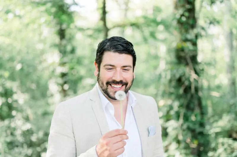 The smiling groom-to-be holding a dandelion clock among the greenery