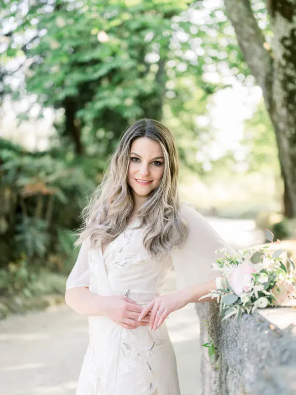 Smiling fiancée on a sunny path, bouquet resting on the stone