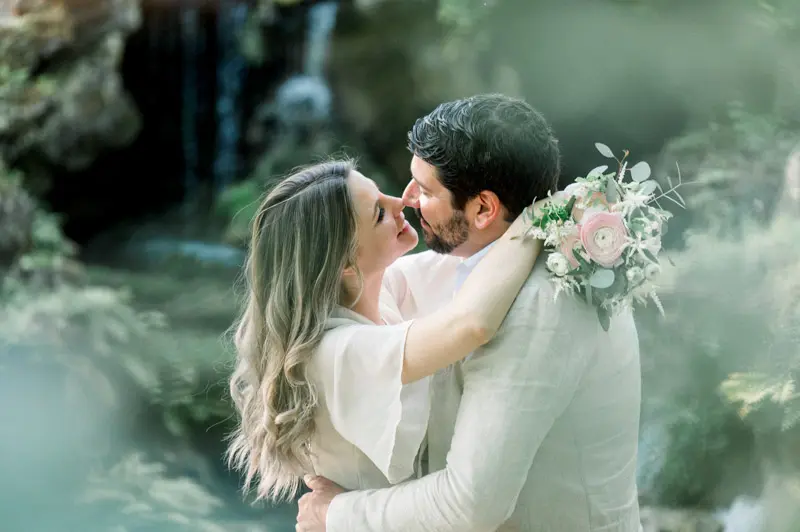 The couple kissing by the waterfall, bouquet resting on Julien's shoulder