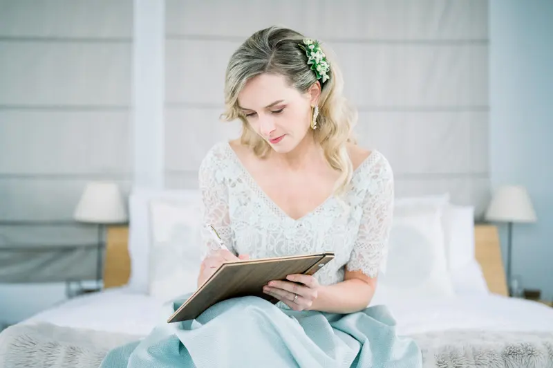 Mariée en robe de dentelle écrivant dans un carnet, assise sur le lit, à Lanton
