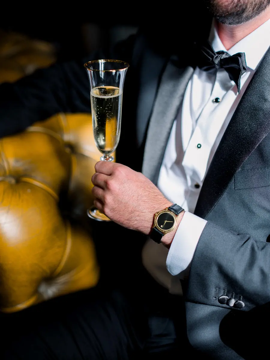 Marié en smoking tenant une flûte de champagne, assis sur un canapé en cuir
