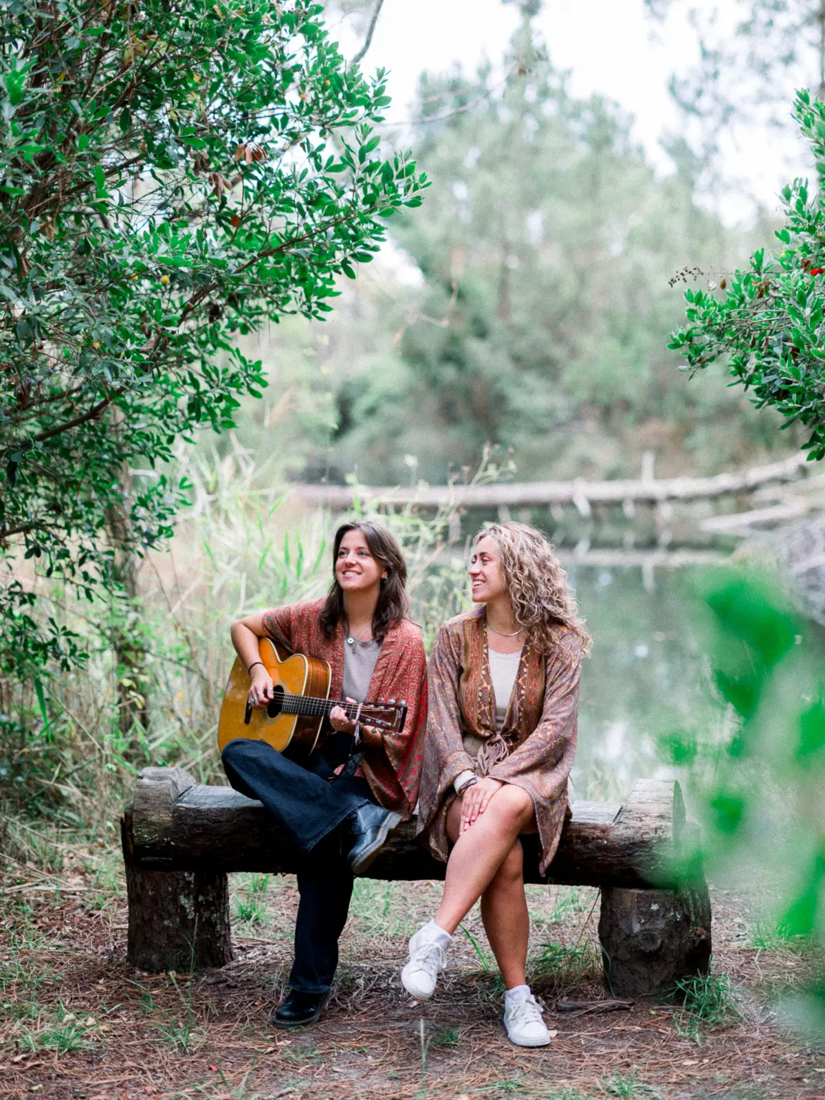 Two women on a wooden bench by a pond, one playing the guitar