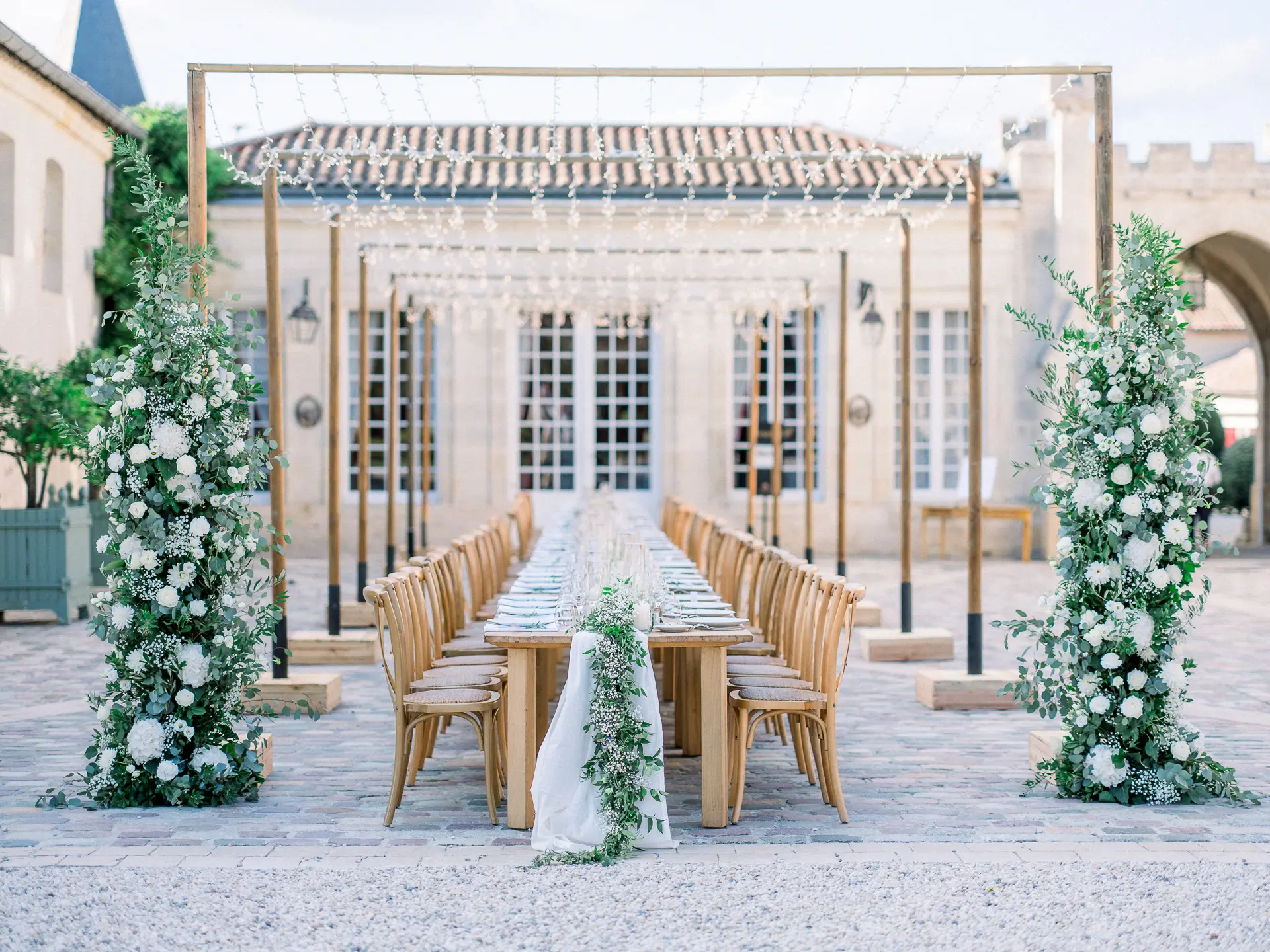 Longue table en bois entre deux arches fleuries devant un château