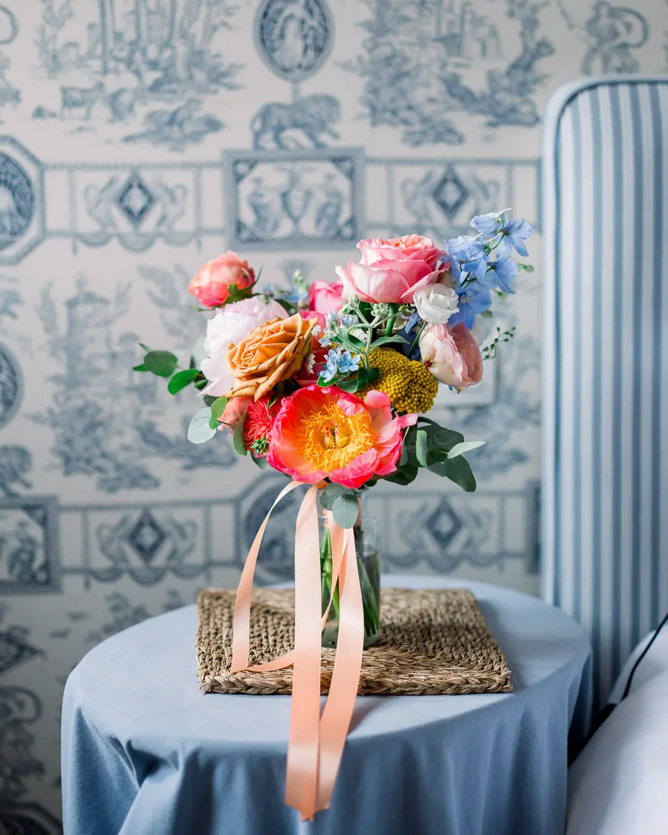 Bouquet coloré sur une table nappée de bleu, devant un papier peint toile de Jouy