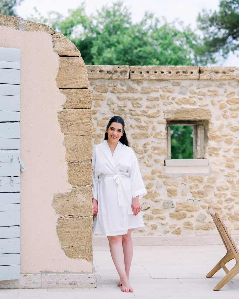Mariée en peignoir blanc, pieds nus, devant un mur en pierre baigné de lumière naturelle